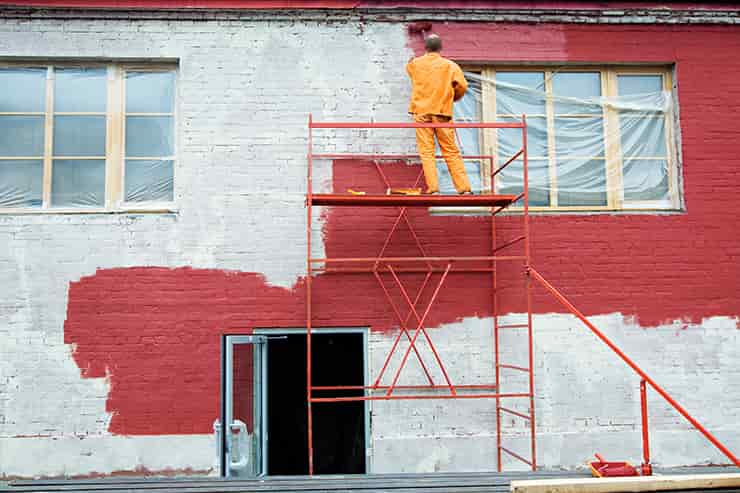 A man on a ladder painting a red brick building, showcasing his skill in building restoration.