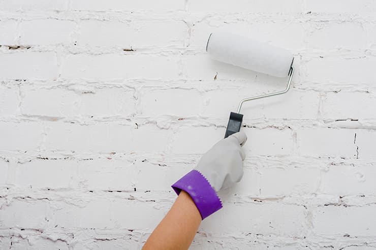 Individual painting a white brick wall with a roller, demonstrating a home improvement project.