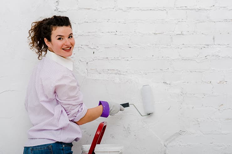 A woman using a paint roller to apply white paint to a wall, engaged in a brick painting activity.