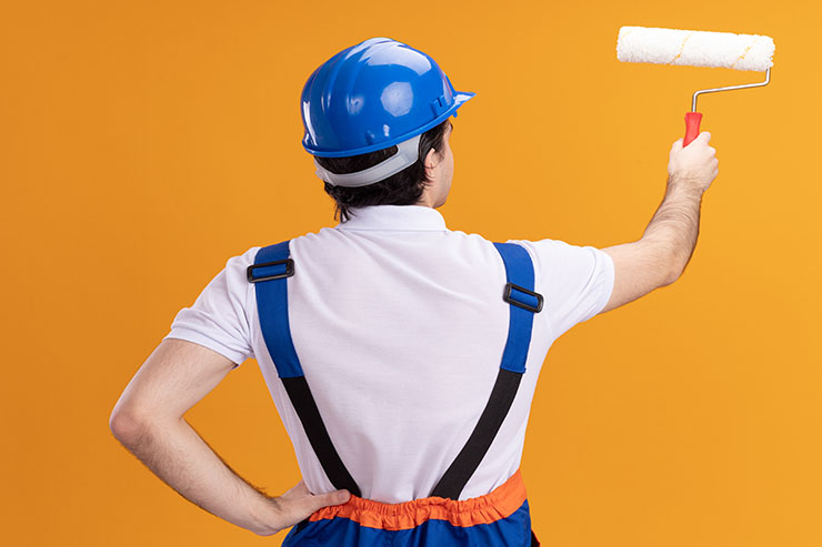 A man wearing overalls and a hard hat stands with a paint roller, ready to start repainting a rental property.