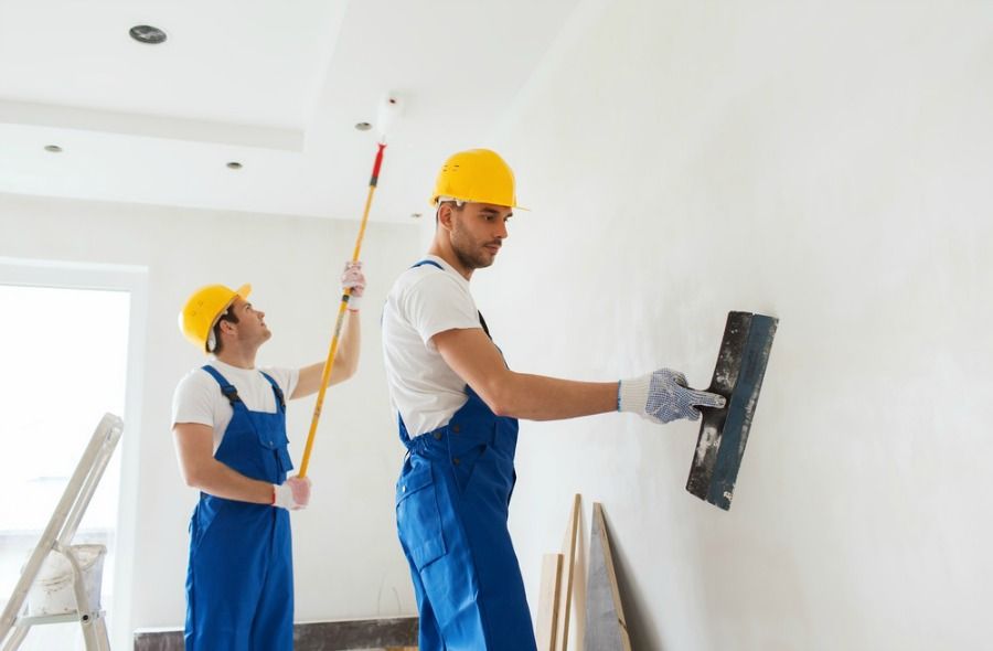Two men in blue overalls and hard hats are painting a wall together with brushes and paint cans nearby.